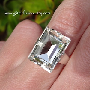 May include: A clear, faceted, rectangular crystal ring set in a silver band. The ring is worn on a finger.