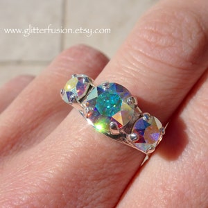 May include: A silver-toned ring featuring three round, iridescent, multi-colored faceted stones set in prongs. The central stone is larger than the two flanking stones, displaying shifting hues of blue, green, and gold. Text reads www.glitterfusion.etsy.com.