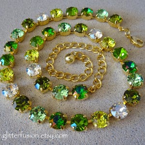 Puede incluir: Un collar dorado con diseño en espiral, con piedras de cristal verdes y transparentes. El collar tiene una cadena y un cierre dorados. Los cristales varían en tonos verdes, creando un efecto degradado. El sitio web www.glitterfusion.etsy.com es visible.