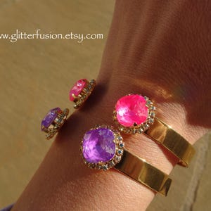 May include: Two gold bangle bracelets with a purple and pink jeweled accent. The bracelets are on a person's wrist.