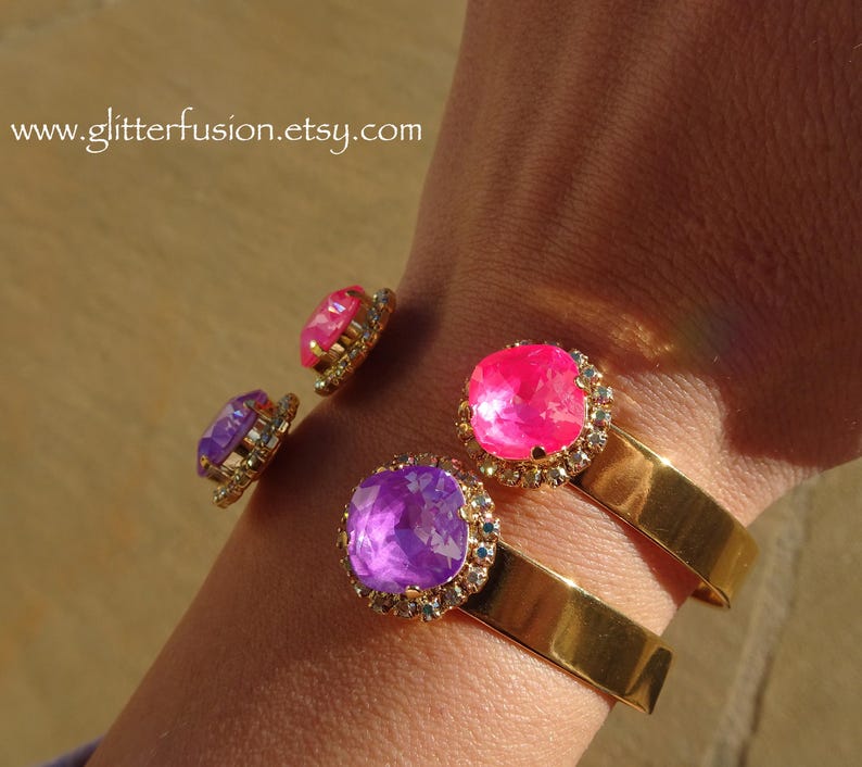 May include: Two gold bangle bracelets with a purple and pink jeweled accent. The bracelets are on a person's wrist.