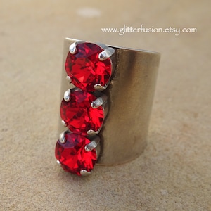 May include: A silver ring with three red rhinestones set in a vertical row. The ring is wide and has a flat band.