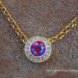 May include: A gold chain necklace with a round pendant featuring a large red and purple gemstone surrounded by smaller clear rhinestones.