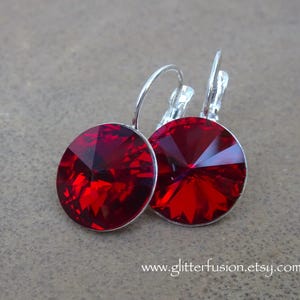 May include: A pair of leverback earrings featuring large, faceted, round, ruby-red crystals. The earrings have silver-colored metal settings and clasps. The crystals reflect light, creating a sparkling effect. The earrings are displayed on a neutral background.