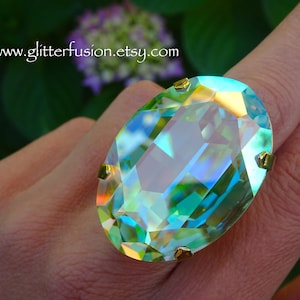 May include: An oval-shaped, iridescent crystal ring featuring a large, faceted stone with light-reflecting green, blue, and yellow hues, secured by four gold-toned prongs. The text www.glitterfusion.etsy.com is visible in the upper left corner.