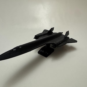 Puede incluir: Un modelo de avión SR-71 negro. El modelo tiene un cuerpo largo y delgado con alas y colas distintivas. El texto "SR-71" es visible en la parte inferior del avión. El modelo está sobre un fondo blanco.