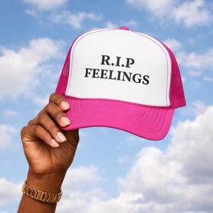 Peut inclure: Casquette de camionneur rose et blanche avec le texte "R.I.P FEELINGS" en noir. Le chapeau est tenu contre un ciel bleu nuageux. La casquette a un bord rose et un dos en filet.