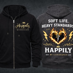 Puede incluir: Sudadera con capucha negra con cremallera y texto dorado &laquo;Happily by Constance&raquo; y una corona. La parte trasera de la sudadera gris oscuro presenta un coraz&oacute;n roto en llamas, rayos y las palabras &laquo;Soft Life, Heavy Standards&raquo;.