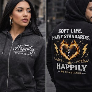 Puede incluir: Sudadera con capucha con cremallera gris carb&oacute;n con la palabra "Happily" en cursiva y el texto "BY CONSTANCE" debajo. La parte trasera de una sudadera con capucha negra presenta un dise&ntilde;o de coraz&oacute;n roto con llamas y rayos, y el texto "SOFT LIFE. HEAVY STANDARDS."
