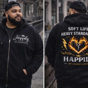 Puede incluir: Sudadera con capucha negra con cremallera con el texto "Happily by Constance" en la parte delantera. La parte trasera presenta un gr&aacute;fico de un coraz&oacute;n en llamas con el texto "Soft Life, Heavy Standards" y "Happily Est. By Constance 2026".