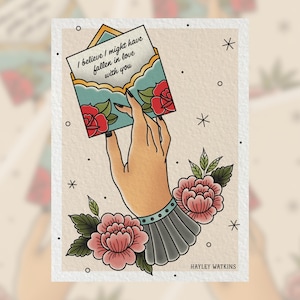 May include: A hand holding a blue envelope with a handwritten message that reads "I believe I might have fallen in love with you". The hand is adorned with a gray cuff and surrounded by pink flowers.