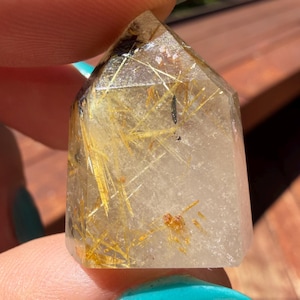 以下が含まれることがあります： 透明感のあるボディに針状の金色のルチルインクルージョンと小さな暗い鉱物の斑点が多数含まれた、磨かれたルチルクォーツのクリスタルポイント。五角形の塔のような形状で、先端が尖ったデザインになっています。