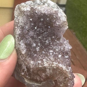 May include: A raw amethyst geode cluster. The geode features a dark purple interior with sparkling crystal formations. The exterior is a rough, brownish-gray rock. The geode is held in a hand with a light green nail polish.