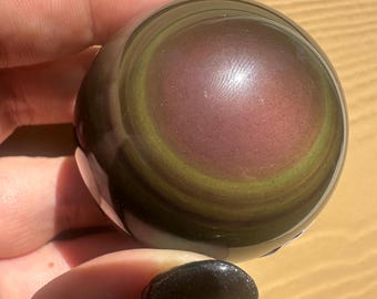 May include: A polished, spherical gemstone with swirling bands of color. The orb features a deep burgundy center, encircled by bands of olive green and dark brown. The stone is held in a hand, showcasing its smooth, reflective surface. A decorative item.