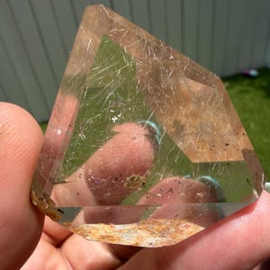 Puede incluir: Un cristal de cuarzo rutilado facetado y transparente que presenta finas inclusiones doradas en forma de aguja por toda la piedra. La base del cristal muestra un depósito mineral rugoso, texturizado y de color marrón anaranjado, mientras que la parte superior es transparente.