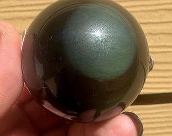 May include: A polished, spherical obsidian gemstone. The orb has a dark, glossy surface with iridescent green and black swirls. The stone is held in a hand, showcasing its smooth, reflective qualities. The gemstone is a natural, decorative item.