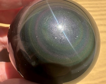 May include: A polished obsidian sphere, displaying iridescent bands of green, purple, and black. The gemstone ball reflects light, creating a starburst effect. The sphere is held in a hand, showcasing its smooth, rounded form.