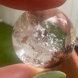 May include: A clear quartz crystal sphere with internal inclusions of brown and reddish-brown minerals. The sphere is held between two fingers with green nail polish.