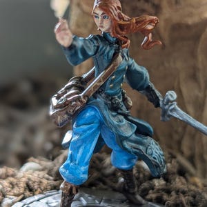 May include: A painted miniature figurine of a female character wearing a blue coat and pants, holding a sword and a brown bag. The figurine is standing on a circular base with a stone pattern.