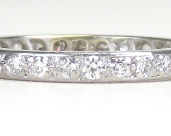 Könnte beinhalten: Ein Platin-Eternity-Ring mit einer durchgehenden Reihe von rund geschliffenen Diamanten. Die Diamanten sind eng aneinander gesetzt und bilden ein funkelndes Band aus weißen Edelsteinen. Der Ring wird auf einer weißen Oberfläche präsentiert.