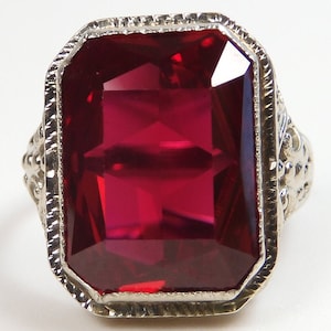 May include: A ring with a large, deep red, emerald-cut gemstone set in a white metal band with intricate detailing.