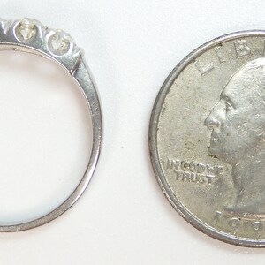 Puede incluir: Una banda de plata con cinco peque&ntilde;os diamantes engastados en fila. El anillo est&aacute; al lado de una moneda de veinticinco centavos de d&oacute;lar estadounidense para comparar el tama&ntilde;o.