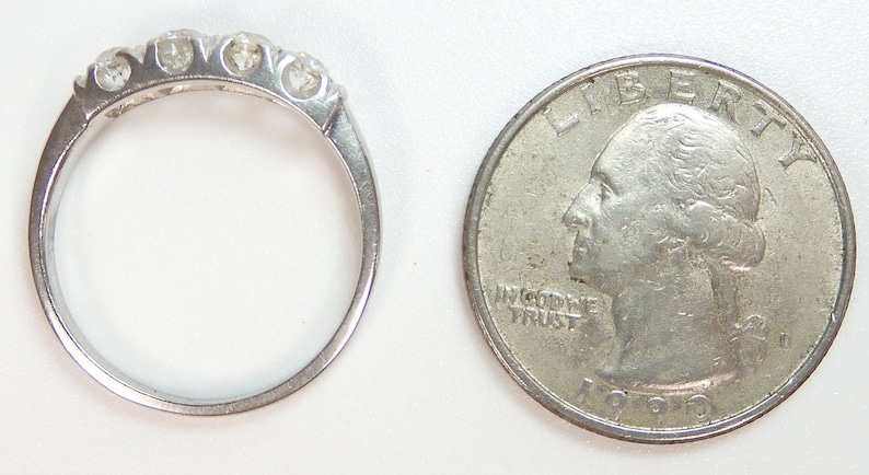 Puede incluir: Una banda de plata con cinco peque&ntilde;os diamantes engastados en fila. El anillo est&aacute; al lado de una moneda de veinticinco centavos de d&oacute;lar estadounidense para comparar el tama&ntilde;o.
