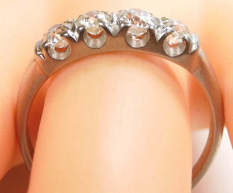 Puede incluir: Un anillo de banda de plata con cinco diamantes redondos engastados en garras. El anillo est&aacute; en un dedo.
