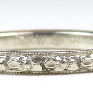 May include: A silver ring with a floral pattern. The ring features a repeating design of stylized flowers around the band. The ring is set against a white background.