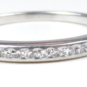 May include: A silver band ring with a row of small, clear gemstones set across the top. The ring is simple and elegant, with a polished finish. The gemstones sparkle against the silver.