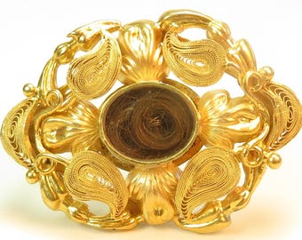 May include: A gold-coloured filigree brooch with a circular glass locket in the centre. The locket contains a lock of hair. The brooch is decorated with paisley-shaped designs.