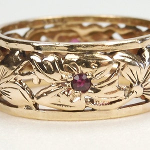 May include: A gold-colored ring with a floral design. The ring features a series of carved flowers and leaves around the band, with a small, round, red gemstone set in the center of some of the flowers. The ring has a vintage or antique appearance.