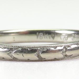 May include: A silver-colored ring with a repeating leaf pattern around the band. The ring has a smooth, polished finish and is likely made of a precious metal. The ring is a piece of jewelry.
