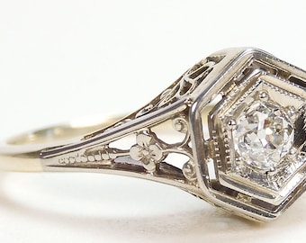 May include: A vintage engagement ring with a hexagonal setting and a single round diamond. The ring is made of white gold and features intricate filigree details.