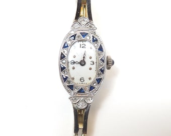 May include: An oval-shaped wristwatch featuring a white dial with black numerals at 3, 6, 9, and 12. The bezel is decorated with triangular blue stones and clear stones set in a silver-toned metal frame. A small crown with a dark accent sits on the side.