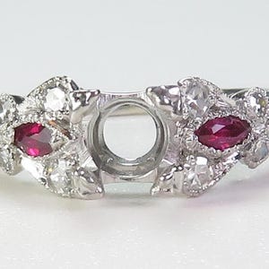 May include: A silver ring with a centre setting for a large gemstone. The ring is decorated with smaller gemstones, including two red rubies.