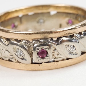 May include: A two-tone ring with a gold band and a silver-colored center band. The center band features a repeating pattern of diamonds and red gemstones. The ring has an intricate, detailed design.