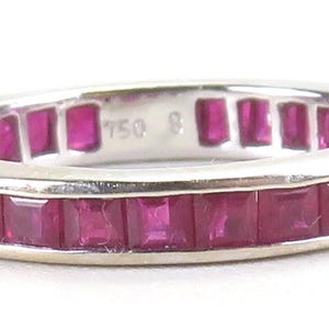 May include: A silver ring with a band of square-cut, vibrant red gemstones. The ring is marked with "750" and "8" on the inside. The gemstones are set closely together, creating a continuous line of color around the ring.