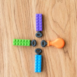 May include: An overhead shot of colorful toy building blocks and key-shaped accessories arranged on a wooden surface. The blocks are green, blue, and purple, with an orange accessory. The arrangement resembles a cross.