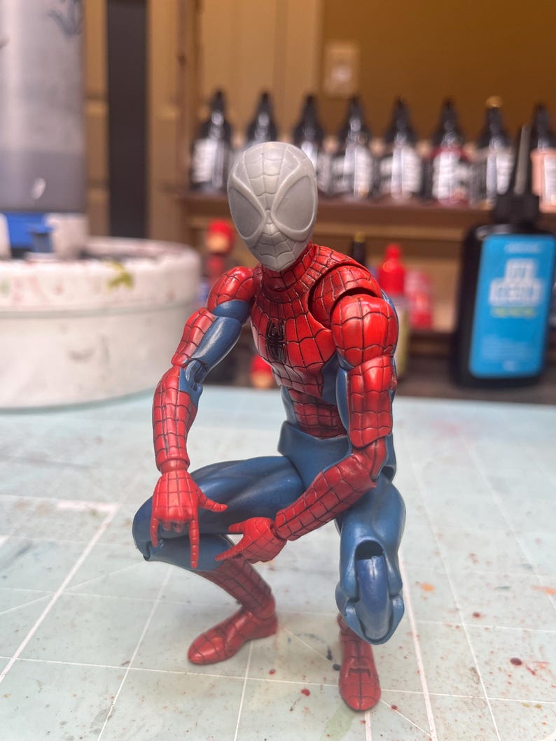 May include: A red and blue action figure of Spider-Man in a crouching pose. The figure has a gray, unpainted head with a spider design.