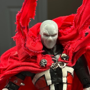 May include: A Spawn action figure featuring a light grey mask, a black and red suit, and a large, tattered red fabric cape. The cape is secured at the front with two silver-coloured skull-shaped clasps connected by a small metal chain link.