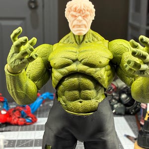 May include: A muscular action figure with green skin, a textured torso, and raised arms. The figure has a light-colored, unpainted head with a grimacing expression and wears black pants with visible distressed holes at the knees and lower legs.