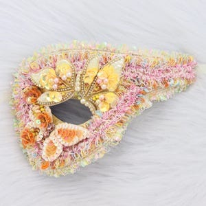 May include: A decorative mask with a butterfly theme. The mask features a central eye opening surrounded by a textured, multi-colored fabric. Embellishments include sequins, beads, and butterfly-shaped appliqués in shades of yellow, orange, and pink.