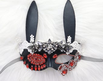 May include: Black masquerade mask with bunny ears, decorated with red and grey rhinestones and white lace. The mask has a half-face design with a black ribbon tie, ideal for a costume party or special event.