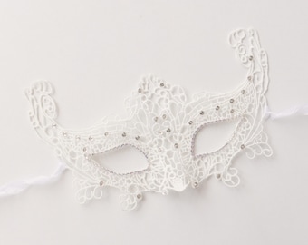 May include: A white lace masquerade mask with rhinestone embellishments. The mask has a detailed floral pattern and eye cutouts. A white ribbon is attached for tying. Ideal for a ball or costume event.