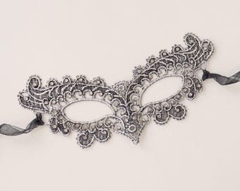May include: A silver masquerade mask crafted from intricate lace with a metallic finish. The mask features ornate swirling patterns and is embellished with small, clear rhinestones throughout. It includes thin black ribbons on each side.