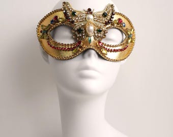 Può includere: Una maschera da ballo dorata con un motivo centrale a farfalla decorato con strass trasparenti, rossi, verdi e blu e una perla bianca centrale. La maschera ha una finitura dorata testurizzata ed è decorata con file di piccoli cristalli rossi.