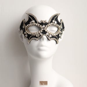 Op de afbeelding: Een vleermuisvormig masker voor een gemaskerd bal, bedekt met zwarte glitters en versierd met heldere, gefacetteerde kristallen van verschillende groottes. Het masker heeft een puntig vleugelontwerp en wordt getoond op een glad, wit etalagehoofd.