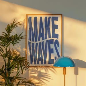 May include: A framed poster with the text "MAKE WAVES" in blue letters on a white background. The poster is hanging on a white wall with a blue lamp in the foreground.