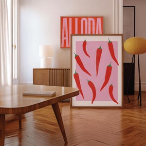 May include: A framed print with a pink background and red chili peppers. The print is hanging on a wall in a living room with a wooden table and a lamp.